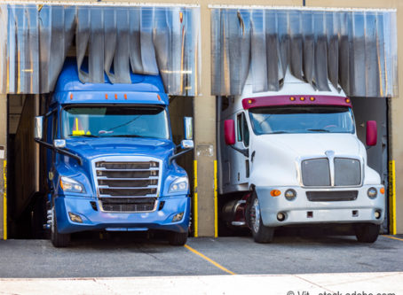 Row of big rigs semi trucks standing inside of warehouse loading commercial cargo. Photo by Vit